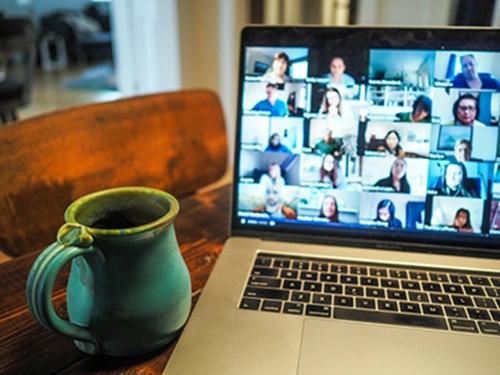 A mug and a laptop computer sitting next to each other on a table. A virtual meeting is taking place on the laptop, which is displaying multiple participants.
