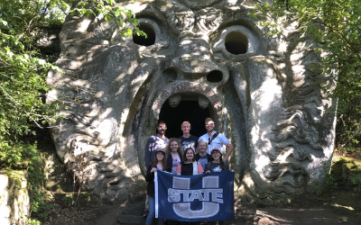 At the Orcus mouth in the Gardens of Bomarzo, Alumnus Helen Lea and other alumni hold a USU Flag