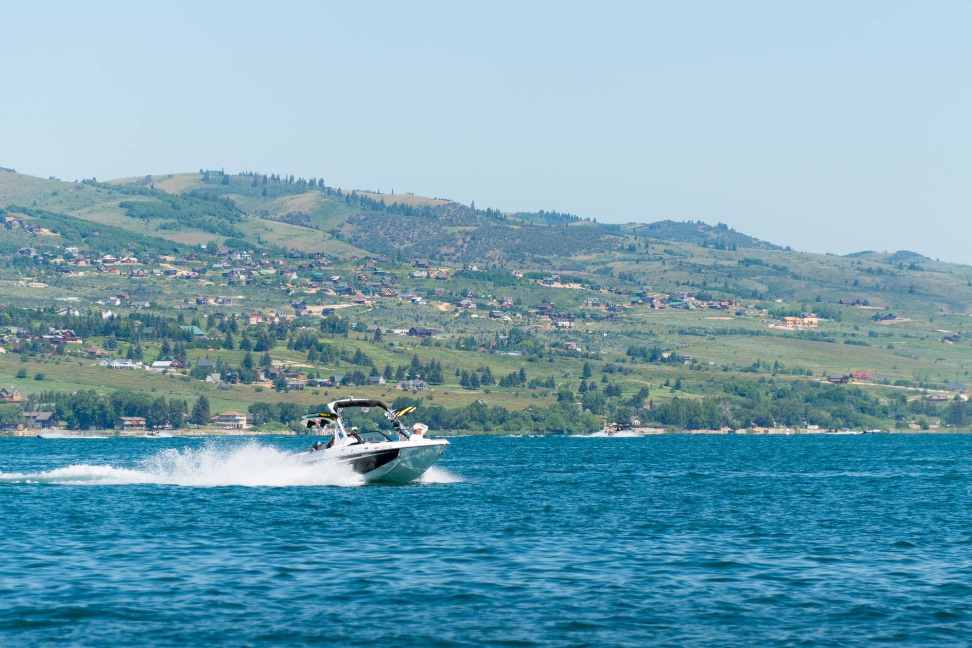 A motorboat sprays water and churns up a wake as it speeds across the surface of Bear Lake on a sunny day. Mountain foothills dotted with cabins can be seen in the background beneath a blue sky.