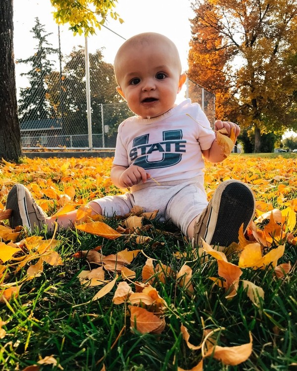 A baby wearing a USU shirt plays with leaves in the grass.