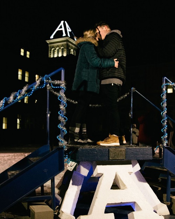 Two students kiss on top of the Block A in front of an illuminated Old Main at midnight.